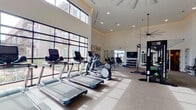 This modern fitness center is designed to support a comprehensive workout experience across multiple dedicated zones. The main exercise floor commands attention with its soaring double-height ceilings, expansive floor-to-ceiling grid-pane windows, and recessed lighting accented by subtle cove illumination. Natural light pours in generously, creating an open and energizing atmosphere. This primary area is outfitted with a full lineup of cardio equipment, including treadmills, elliptical trainers, and a stair climber, alongside a cable-based functional training rig, a flat-screen television, and a rack of kettlebells. A separate free weights room features wood-look tile flooring, full-wall mirrors, a comprehensive dumbbell rack, and an assortment of exercise balls and resistance tools, providing ample space for strength and flexibility training. A dedicated spin studio offers a focused cycling experience, with a row of screen-equipped stationary bikes, mirrored walls, and framed wall art enhancing the boutique-style ambiance. Additional areas include a strength machine zone and accessory spaces with proper ventilation and clean finishes throughout. The overall design balances a bright, contemporary aesthetic with practical functionality, making this fitness center a well-rounded facility suited for a wide range of fitness routines.