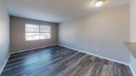 This single-floor unit offers a clean, modern aesthetic with thoughtfully updated finishes throughout. The living area is open and welcoming, featuring durable wood-style laminate flooring in a cool gray tone that lends a contemporary feel to the space. A standout architectural detail is the pass-through window connecting the living area to the kitchen, complete with a countertop overhang that functions as a convenient breakfast bar, enhancing the sense of openness between the two spaces.

The kitchen is efficiently designed with white shaker-style cabinetry, granite-look countertops, a stainless steel single-basin sink, and a stainless steel gas range with an overhead exhaust hood — offering a functional setup for everyday cooking.

The bedroom features plush carpeting for added comfort and includes a wide sliding-door closet that provides generous storage capacity. A second room, also with wood-style flooring, benefits from natural light streaming through a window fitted with blinds, making it a versatile space suitable as a bedroom or home office.

The bathroom is neatly appointed with wood-look flooring, a white vanity with mirror cabinet, and a full tub-shower combination with tiled surround. Throughout the unit, neutral wall tones and clean white trim create a bright, cohesive atmosphere that is move-in ready.