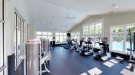 This fitness center is housed in a single-story structure designed to maximize both functionality and comfort. The space features vaulted ceilings accented by a centrally mounted ceiling fan and recessed lighting, contributing to an open, airy atmosphere. Large, white-trimmed windows line multiple walls, flooding the interior with abundant natural light and offering views of surrounding greenery and what appears to be a pool area beyond the glass. The dark rubber flooring provides a durable, slip-resistant surface well-suited to high-traffic exercise use. The equipment layout is thoughtfully organized, with a row of cardio machines — including treadmills, ellipticals, and stationary bikes — positioned to take advantage of the window views. A cable strength training machine is prominently featured on one side, rounding out the facility's offerings. The soft, neutral wall tones complement the crisp white window trim, creating a clean, professional aesthetic. A wall-mounted television adds convenience for users during workouts. Overall, the space strikes a balance between a bright, welcoming environment and a fully functional fitness facility.