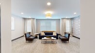 Located at 401 Lime Quarry Road in Madison, Alabama, this single-floor space offers a thoughtfully designed communal environment with a clean, contemporary aesthetic. The central lounge area serves as the focal point, featuring a navy blue tufted sofa paired with rich brown leather armchairs arranged around a sleek metal-framed coffee table. Recessed lighting and a central pendant fixture illuminate the room, while multiple windows dressed with white horizontal blinds and sheer linen drapes allow natural light to filter throughout.

A distinctive accent wall covered in a soft, wave-patterned wallcovering in muted blue and gray tones adds visual interest to one seating alcove, complementing the neutral gray walls and warm-toned carpet that flows consistently throughout the space. The carpeting provides a cohesive, comfortable foundation across the lounge and connecting corridors.

Wide hallways extend from the lounge area, lined with pendant lighting and dark wood baseboards that offer a refined contrast to the light-toned walls. Elevator access is conveniently positioned along the corridor, suggesting this space is part of a larger multi-story building. The overall ambiance is calm, welcoming, and professionally appointed, reflecting a well-maintained communal or hospitality-oriented environment.