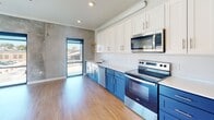 This single-floor space showcases a contemporary design with a cohesive blend of modern finishes and industrial-inspired details. Warm hardwood flooring runs throughout, connecting each area seamlessly and contributing to the open, airy feel of the layout.

The open-concept living and kitchen area serves as the heart of the space. Large, black-framed sliding glass doors and windows flood the room with natural light and provide direct views of the surrounding streetscape and greenery. A striking exposed concrete accent wall adds an industrial character that contrasts effectively with the crisp white walls and recessed lighting overhead. Built-in floating wood shelves offer both functional storage and a decorative element to the living area, complemented by a wall-mounted mini-split system for climate control.

The kitchen is a standout feature, equipped with bold cobalt blue lower cabinets paired with white upper shaker-style cabinetry, creating a vibrant two-tone aesthetic. Live-edge wood open shelving adds a natural warmth, while stainless steel appliances—including a range, microwave, dishwasher, and refrigerator—provide full functionality on white countertops.

A well-proportioned hallway leads to additional rooms, including a bathroom outfitted with a full tub-shower combination, wood-toned vanity, and clean tile flooring. The bedroom features large windows with outdoor views and a mini-split unit.