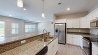This two-story home offers a clean, modern design with a neutral palette and quality finishes throughout. The main level centers around an open-concept layout where the kitchen flows seamlessly into the living area, creating a spacious and connected feel. The kitchen is well-equipped with white shaker-style cabinetry, granite countertops, a full suite of stainless steel appliances including a refrigerator, electric range, and built-in microwave, and a large center island with a double undermount sink and pendant lighting above. Light wood-look flooring runs throughout the main living areas, complementing the bright, airy atmosphere enhanced by recessed lighting.

The upper level, as seen in the dollhouse view, includes bedrooms finished with plush carpeting, along with bathrooms featuring granite-topped white vanities and floor-to-ceiling tile work. One bathroom includes a walk-in glass-enclosed shower with stone-look tile surround, while another features a tiled soaking tub. A dedicated laundry room provides added convenience, complete with a utility sink and upper shelving. Throughout the home, consistent use of granite, white cabinetry, and neutral tones creates a cohesive and polished aesthetic.