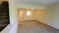 This two-floor residential space offers a clean, functional layout designed for comfortable everyday living. The interior is finished with neutral, light-toned walls throughout, creating a bright and welcoming atmosphere that feels open and airy.

The main living area is generously sized, featuring wall-to-wall carpeting in a warm beige tone that adds softness underfoot. The room benefits from natural light through a window, and a flush-mount ceiling light provides additional illumination. Subtle architectural detailing, including a partial soffit or beam element near the ceiling, adds a touch of dimension to the otherwise streamlined space.

A wide open entryway connects the main living area to an adjacent corridor and additional rooms beyond, suggesting a practical and well-considered floor plan. The hallway leads further into the unit, hinting at additional living spaces, storage, or utility areas on the same level. The second floor expands the overall footprint of the space, offering additional rooms and living flexibility.

Throughout, the consistent use of neutral carpeting, clean white trim, and pale wall finishes gives the space a cohesive, move-in-ready aesthetic. The overall ambiance is understated and versatile, providing a neutral canvas well-suited to a variety of furnishing styles and personal touches.