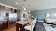 Located at 1715 Lake Michigan Dr in Valparaiso, Indiana, this well-appointed single-floor residence offers a thoughtfully designed open-concept layout with quality finishes throughout. The kitchen serves as a central hub, showcasing rich dark wood cabinetry, granite countertops, and a large island with an integrated sink, dishwasher, and bar seating. Stainless steel appliances — including a gas range, microwave, and French door refrigerator — complement the warm hardwood flooring that flows seamlessly into the dining and living areas. The dining space sits adjacent to the kitchen and features a dedicated area with access to a hallway leading to the private quarters. The living room is bright and welcoming, with large windows that invite abundant natural light and views of the surrounding neighborhood, accented by cool blue-gray walls and a ceiling fan. The bedroom offers plush carpeting, natural light from a window, and generous space for furnishings. The bathroom is finished with granite vanity countertops, dark wood cabinetry, hardwood-style flooring, and a glass-enclosed walk-in shower with tile surround. A dedicated utility room houses an in-unit washer and dryer alongside the water heater and HVAC system, providing added convenience.