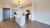 This well-appointed single-floor residence offers a thoughtfully designed layout that balances comfort and modern style. The open-concept kitchen serves as a central highlight, showcasing crisp white cabinetry, a clean white countertop with a peninsula island, and a full suite of stainless steel appliances including a gas range, built-in microwave, dishwasher, and French door refrigerator. A pendant chandelier above the island and track lighting overhead provide layered illumination, while wood-look luxury vinyl plank flooring ties the kitchen and living areas together seamlessly. The living area benefits from an abundance of natural light, with a large sliding glass door opening to a private balcony that offers open outdoor views of the surrounding area. The bedroom is carpeted for comfort and features a generous window that frames outdoor views and fills the room with natural light. The bathroom is finished with large-format tile flooring, white cabinetry with dark hardware, a combined tub and shower, and clean white fixtures throughout. The overall design palette of warm neutrals, crisp whites, and contemporary finishes gives the space a bright, move-in-ready ambiance suited to modern living.
