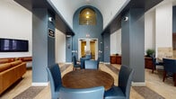 This single-floor community space is thoughtfully designed to accommodate a variety of social and functional needs. The entryway immediately impresses with its soaring vaulted ceiling and a striking arched architectural detail finished in deep blue-gray tones, framing the entrance and lending the space a grand, welcoming character. Tile flooring in warm neutral tones flows throughout, unifying the distinct zones within the layout.

The lounge area features soaring white walls that amplify the sense of openness, furnished with contemporary blue accent chairs, an orange sofa, a wall-mounted flat-screen television, and a full-length shuffleboard table, creating an inviting setting for casual recreation and socializing. Adjacent to the lounge, a dining area is anchored by a round wood table surrounded by coordinating blue upholstered chairs, reinforcing the cohesive color palette.

The fully equipped kitchen is well-appointed with rich dark wood cabinetry, granite countertops, a travertine tile backsplash, and a complete suite of stainless steel appliances including a side-by-side refrigerator, double wall oven, and dishwasher. A separate game room visible in the floor plan includes a pool table, rounding out this multifunctional communal facility.