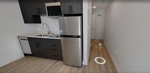 This 200 square foot studio apartment in Salt Lake City offers a thoughtful blend of functionality and style. The single-floor layout comprises a modern kitchen and a full bathroom, efficiently utilizing every inch of space. The kitchen boasts sleek dark wood cabinetry, stainless steel appliances including a refrigerator and built-in microwave, and a subway tile backsplash. A sink with a modern faucet and ample counter space complete the culinary area. The bathroom features a walk-in shower with a glass partition, a standard toilet, and necessary fixtures like a towel ring and toilet paper holder. Light wood-look flooring runs throughout the apartment, creating a sense of continuity and openness. A large floor-to-ceiling window floods the main living area with natural light, enhancing the sense of space and providing a connection to the outdoors. The neutral color palette, dominated by whites and grays, contributes to the apartment's contemporary ambiance. Despite its compact size, the layout appears well-planned, offering a comfortable living space for urban dwellers.