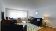 Located at Westgate, this single-floor space offers a comfortable and stylishly arranged living area that balances practicality with design appeal. The living room serves as the centerpiece of the space, featuring warm hardwood flooring that anchors the room and adds a sense of natural warmth throughout. A large window dressed with sheer white curtains allows generous natural light to filter in, creating a bright and airy atmosphere. A wall-mounted air conditioning unit provides climate control directly within the room. The space is furnished with a deep navy blue sofa accented by decorative throw pillows in complementary patterns, accompanied by coordinating armchairs that create a cohesive and inviting seating arrangement. A glass-top coffee table sits atop a bold green and white geometric-patterned area rug, adding a contemporary focal point to the room. A starburst mirror mounted on the wall serves as an elegant decorative accent, enhancing the room's visual depth. Soft, warm lighting from table lamps contributes to a relaxed ambiance. Overall, the space presents a clean, well-maintained interior with a thoughtful balance of comfort and style.