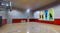 This indoor basketball court is a well-appointed athletic facility designed for serious recreational play and training. The space features a gleaming, polished hardwood floor with clearly marked court lines, including a three-point arc and key, providing a professional playing surface. Regulation-height basketball hoops are mounted on opposing ends of the court, offering a full game-ready setup. The walls are finished in a two-tone design, with a bold red lower half and a neutral gray upper half, creating an energetic and sporty atmosphere. A striking mural on one wall depicts silhouetted athletes in dynamic poses against vibrant color blocks of red, yellow, and blue, adding a lively visual focal point to the space. The court is illuminated by bright overhead lighting that ensures excellent visibility throughout the playing area. Adequate ceiling height allows for unobstructed play, including full shooting arcs and elevated passes. Ventilation units are present along the lower walls, maintaining air circulation and comfort during active use. A secured door provides access to and from the court. Overall, the space combines functionality with an inspiring aesthetic, making it an excellent venue for basketball and general athletic activities.