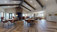 Located in Waunakee, Wisconsin, this single-floor community clubhouse on Connery Cove is a thoughtfully designed gathering space that blends rustic warmth with modern sophistication. The main hall immediately impresses with soaring vaulted ceilings accented by bold dark wood exposed beams and a striking herringbone-patterned hardwood floor that flows throughout. A floor-to-ceiling stacked stone fireplace anchors the lounge area, where eclectic, colorful armchairs and warm lighting create an inviting atmosphere. French doors adjacent to the fireplace open to an outdoor area, bringing in natural light and a seamless indoor-outdoor connection. The full kitchen is equipped with stainless steel appliances, a professional-grade range with hood, light cabinetry, and generous counter space, all illuminated by large black-framed windows overlooking open green surroundings. Multiple dining zones feature round wood tables with rattan and wire-frame chairs, complemented by high-top bistro tables adorned with vibrant landscape artwork above. A quieter lounge nook offers plush red armchairs, a tufted leather ottoman, and a statement gold chandelier, ideal for intimate conversations. A decorative wall display of woven baskets adds an artful, eclectic touch. Throughout, the space maintains an energetic yet comfortable ambiance suited for both casual gatherings and community events.