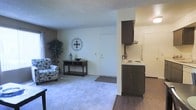 This single-floor residential space is designed with an open-concept layout that seamlessly connects the living area and kitchen, creating a functional and cohesive environment. The living area features plush carpeted flooring and neutral-toned walls that lend a clean and understated aesthetic, complemented by a textured ceiling that adds a classic touch. The space is naturally brightened by light-colored walls, giving the room an airy, welcoming feel. A decorative wall accent adds a subtle artistic detail, while the room comfortably accommodates seating and accent furniture. Adjacent to the living area, the kitchen transitions to hard surface flooring and is equipped with dark wood cabinetry that provides a warm contrast to the lighter surroundings. The kitchen includes a countertop range and under-cabinet range hood, with additional cabinetry above for ample storage. A side-by-side refrigerator completes the kitchen setup. The entry door is conveniently located at the far end of the living space, making for a straightforward and practical layout. Overall, this space offers a compact yet well-organized floor plan suited for comfortable everyday living.