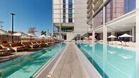 This exceptional outdoor amenity level is set at the base of a contemporary high-rise tower, showcasing a sophisticated architectural design defined by exposed concrete columns, expansive floor-to-ceiling glass facades, and clean geometric lines. The centerpiece of the space is a pair of generously sized, crystal-clear lap pools that run parallel to one another, separated by a wide tiled walkway. The vivid turquoise water reflects the surrounding structure and open sky, lending the area a resort-like ambiance. On one side of the pool, rows of cushioned lounge chairs with warm orange upholstery are arranged beneath white shade umbrellas, offering a comfortable setting for relaxation. The opposite side features additional seating nestled beneath a covered colonnade, providing a shaded retreat. A vibrant large-scale mural adds a bold artistic focal point to the covered indoor-outdoor lounge area, which appears to include additional seating and bar amenities. An open-air staircase connects levels within the structure, adding a dynamic vertical element. The deck opens to panoramic views of the surrounding cityscape and distant mountain ranges, bathed in abundant natural sunlight. The overall atmosphere is modern, energetic, and polished, combining urban sophistication with relaxed outdoor living.