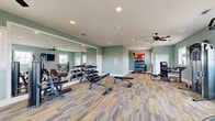 This fitness center offers a generously sized, single-floor workout facility designed to accommodate a wide range of exercise needs. The space is finished with soft sage-green walls that create a calm, focused atmosphere, complemented by multi-toned patterned carpet flooring throughout most of the floor area, with wood-look flooring visible in the cardio zone.

Natural light fills the room through multiple large windows along the perimeter walls, providing bright, airy conditions for workouts. Ceiling fans are strategically placed throughout to maintain comfortable airflow, while recessed lighting and overhead HVAC vents ensure year-round climate control.

The cardio section features treadmills, a stair climber, and an elliptical machine, all positioned facing the windows to take advantage of the natural views. A large wall-mounted flat-screen television accompanies the cardio area. The strength training zone includes a cable/pulley machine, a leg press, free-weight benches, and a full dumbbell rack. A dedicated functional training corner houses kettlebells, medicine balls, resistance bands, TRX straps, and stability balls neatly organized on a multi-tier storage rack.

Floor-to-ceiling mirrors along one wall visually expand the space and assist users in monitoring their form. The facility is clean, well-organized, and thoughtfully laid out to support a comprehensive fitness routine.