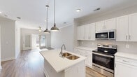 Located at 1025 Hirschfield Dr in Fayetteville, NC, this single-story home offers a thoughtfully designed layout with modern finishes and a bright, open feel throughout. The kitchen serves as the heart of the home, showcasing white shaker-style cabinetry, a subway tile backsplash, light quartz countertops, and a central island with a double stainless steel sink. Stainless steel appliances, including a refrigerator, electric range, microwave, and pendant lighting overhead, complete the space. The kitchen flows seamlessly into the living area, which features warm wood-look flooring, crown molding, and a ceiling fan—a consistent detail found across multiple rooms. Natural light enters generously through windows fitted with white blinds. The primary bedroom is a comfortable retreat with its own walk-in closet featuring wire shelving and an en-suite bathroom equipped with a white vanity and a tub-shower combination. Additional bedrooms are well-sized, each with ceiling fans and wood-look flooring. A secondary bathroom mirrors the clean, neutral aesthetic with matching white fixtures. Walk-in closets with wire shelving provide ample storage throughout. The overall ambiance is clean, contemporary, and move-in ready.