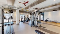 This fitness center offers a comprehensive workout environment designed with both functionality and aesthetics in mind. The space is organized across a single open floor, allowing for a clear distinction between cardio, strength, and functional training zones. Large windows along multiple walls flood the interior with natural light, creating an open, energizing atmosphere while providing outdoor views.

The cardio section features a row of treadmills, elliptical trainers, and upright bikes, all positioned to face the windows. A dedicated strength training area includes a multi-station Octagon cable machine, a full dumbbell rack with weights of varying sizes, adjustable benches, and TRX suspension straps for functional training. A rowing machine occupies a separate transitional area near the entrance corridor.

Architectural details include coffered ceilings with recessed lighting, structural columns finished in white, and bold pendant light fixtures that anchor each zone. Ceiling fans contribute to air circulation throughout. Full-length mirrors along one wall enhance the sense of depth and allow users to monitor their form. The flooring features durable, multi-tone rubber surfacing suited for high-traffic fitness use. Motivational signage near the entrance adds a purposeful, goal-oriented ambiance to the overall environment.