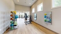 Located at 1101 Club Exchange Dr in Holly Springs, NC, this fitness center offers a generously sized, single-floor exercise room designed to support a full range of workout activities. The space features soaring vaulted ceilings with exposed beam detailing and a white shiplap ceiling finish, giving the room an airy, open feel. Stylish industrial-style pendant lights and ceiling fans are suspended overhead, ensuring excellent air circulation and comfortable conditions throughout workouts.

Natural light flows generously into the space through large, multi-pane windows positioned along multiple walls and at elevated heights, creating a bright and energizing atmosphere. Light-toned hardwood-style flooring runs throughout, adding warmth and a clean aesthetic. Wall-mounted mirrors enhance the sense of depth and allow users to monitor their form.

The exercise room is comprehensively outfitted with a wide selection of equipment, including multiple treadmills, elliptical trainers, stationary and spin bikes, and a stair climber for cardiovascular workouts. Strength training is well supported with selectorized weight machines, a full dumbbell rack, a weight bench, and an assortment of functional fitness accessories such as stability balls, resistance bands, and foam rollers. A wall-mounted television is also present for entertainment during workouts. The overall ambiance is modern, clean, and motivating, making this facility a well-rounded destination for fitness.