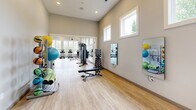 Located at 1101 Club Exchange Dr in Holly Springs, NC, this fitness center offers a generously sized, single-floor exercise room designed to support a full range of workout activities. The space features soaring vaulted ceilings with exposed beam detailing and a white shiplap ceiling finish, giving the room an airy, open feel. Stylish industrial-style pendant lights and ceiling fans are suspended overhead, ensuring excellent air circulation and comfortable conditions throughout workouts.

Natural light flows generously into the space through large, multi-pane windows positioned along multiple walls and at elevated heights, creating a bright and energizing atmosphere. Light-toned hardwood-style flooring runs throughout, adding warmth and a clean aesthetic. Wall-mounted mirrors enhance the sense of depth and allow users to monitor their form.

The exercise room is comprehensively outfitted with a wide selection of equipment, including multiple treadmills, elliptical trainers, stationary and spin bikes, and a stair climber for cardiovascular workouts. Strength training is well supported with selectorized weight machines, a full dumbbell rack, a weight bench, and an assortment of functional fitness accessories such as stability balls, resistance bands, and foam rollers. A wall-mounted television is also present for entertainment during workouts. The overall ambiance is modern, clean, and motivating, making this facility a well-rounded destination for fitness.