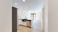 This thoughtfully designed single-floor residence offers a bright, contemporary living experience with a cohesive aesthetic throughout. Light hardwood flooring flows seamlessly across every room, complementing the crisp white walls and creating a clean, airy atmosphere.

The open-concept kitchen is a standout feature, outfitted with white shaker cabinetry, dark countertops, a stainless steel appliance suite including a refrigerator, electric range, and microwave, plus a kitchen island with a matte black farmhouse sink. Globe pendant lights with brass fixtures add a warm, modern touch overhead. The kitchen opens directly into the living area, which benefits from natural light streaming through large, wood-framed windows and a glass door that frames views of the surrounding greenery.

The bathroom is well-appointed with a white vanity, dark countertop, a framed mirror with gold-accented sconce lighting, and a full tub with a sliding glass shower enclosure. In-unit laundry is conveniently included via a stacked front-load washer and dryer. The bedroom features wood-framed windows with transom panels overlooking an outdoor balcony railing and tree-lined scenery. A generously sized walk-in closet with wire shelving provides ample storage. Recessed lighting throughout completes this move-in ready space.
