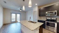This single-floor residence is designed with a clean, modern aesthetic and thoughtful attention to detail throughout. The heart of the home is the open-concept kitchen and living area, where warm hardwood-style flooring flows seamlessly underfoot. The kitchen is outfitted with rich dark cabinetry, a classic tile backsplash, and a full suite of stainless steel appliances, including a refrigerator, range, and built-in microwave. A generous granite-topped island with an undermount sink and elegant pendant lighting above serves as a natural focal point, ideal for both meal preparation and casual dining. A ceiling fan overhead and double French doors allow natural light to fill the living space, creating a bright and welcoming atmosphere. The bedrooms are carpeted for comfort and each benefit from ceiling fans and large windows with exterior views. One bedroom features direct access to a walk-in closet, providing ample storage. The primary bathroom impresses with an L-shaped dual-vanity counter finished in matching granite, dark cabinetry with brushed hardware, dual framed mirrors, and hardwood-style flooring that ties the space together cohesively. The overall ambiance is contemporary and well-finished, with a consistent palette of dark cabinetry, granite surfaces, and neutral tones throughout.