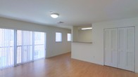 This single-floor unit offers a clean, open layout designed for comfortable everyday living. The main living area is bathed in natural light, courtesy of sliding glass doors that open to a private balcony and additional windows fitted with vertical blinds. The light-toned, hardwood-style flooring runs throughout the space, contributing to a warm and welcoming atmosphere. A half-wall pass-through counter connects the living area to the kitchen, creating an open, communicative flow between spaces while maintaining a sense of defined zones. The kitchen features under-cabinet fluorescent lighting, providing ample task illumination. The neutral white walls and simple flush-mount ceiling light in the living area keep the aesthetic clean and versatile, ready to adapt to a variety of décor styles. A louvered closet door is visible along one wall, offering practical storage solutions. Overall, the space strikes a balance between functionality and brightness, making it an inviting and adaptable environment with a straightforward, modern sensibility.