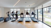 Located on Faulkner Street in Durham, NC, this single-floor fitness center offers a thoughtfully designed layout across multiple connected zones, each tailored to a specific type of training. The space is finished with warm hardwood-style flooring throughout, crisp white walls, and linear LED lighting that runs along the ceilings, creating a clean, contemporary aesthetic.

The cardio zone is lined with floor-to-ceiling windows and large glass panels that flood the area with natural light and provide outdoor views. It is equipped with multiple treadmills, elliptical machines, rowing machines, stair climbers, and stationary bikes, including Assault Fitness models. A wall-mounted television is positioned for use during cardio sessions.

The strength and functional training area features a cable machine system, a full dumbbell rack, barbells, medicine balls, stability balls, a punching bag, and TRX suspension equipment. Full-length mirrors line portions of the walls, enhancing the sense of space. A ceiling fan provides additional air circulation.

A separate studio room, accessible through striking black-framed sliding barn doors, offers open floor space for group classes or individual training, with a ballet barre, additional equipment storage, and natural light from an exterior window. Access to the facility is secured via a resident entry keycard system.