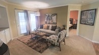 This single-floor residence offers a thoughtfully designed layout with a warm, inviting ambiance throughout. The living area serves as the heart of the space, showcasing neutral earth-toned walls accented by a rich, darker feature wall that adds depth and character. Crown molding runs along the ceiling, lending a refined architectural detail to the room. Plush wall-to-wall carpeting provides comfort underfoot, complemented by layered area rugs that define the seating zone. Natural light flows in generously through large windows dressed with patterned drapes, creating a bright yet cozy atmosphere. The living space is styled with a generous sofa, coordinating accent chairs, and paired ottoman coffee tables, offering both comfort and functionality. A bold aviation-themed wall art piece serves as a striking focal point above the seating arrangement. An open passageway leads directly into the bedroom, which features warm terracotta-toned walls, continuing the cohesive color palette seen throughout the home. The bedroom is furnished with dark wood furniture and offers a sense of privacy while maintaining a visual connection to the main living area. Overall, the space presents a harmonious blend of comfort, style, and practical design.