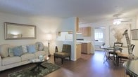 This single-story space is designed with an open-concept layout that seamlessly connects the living area, kitchen, and bedroom, creating a cohesive and functional flow throughout. The living area serves as a welcoming focal point, featuring warm hardwood-style flooring, a comfortable sofa, and an accent chair arranged around a glass-top coffee table. A floor lamp provides warm ambient lighting, complemented by a decorative framed mirror on the wall and a soft area rug that adds texture underfoot.

A partial wall with a pass-through cutout divides the living space from the kitchen, maintaining visual openness while defining each zone. The kitchen is equipped with light wood cabinetry, a white refrigerator, and a dark-toned dishwasher, with a clean countertop providing functional workspace. A range with visible controls sits along the back wall beneath the upper cabinets.

Beyond the kitchen, a glimpse into the bedroom reveals a bright, naturally lit space with a window that invites daylight into the unit. The bedroom accommodates a bed and a small side table, offering a restful retreat. The consistent flooring and neutral color palette throughout the space contribute to a unified, airy ambiance.