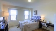 This bedroom offers a clean and inviting atmosphere, characterized by its bright, neutral palette and thoughtful design details. Two double-hung windows along the exterior wall flood the room with natural light, framing views of the surrounding environment while keeping the space feeling open and airy. The windows are fitted with white horizontal blinds, allowing for adjustable light control and privacy.

The flooring is hardwood, adding warmth and a timeless quality to the room. A patterned area rug anchors the sleeping area, introducing subtle texture underfoot. The ceiling is smooth and white, fitted with a decorative flush-mount light fixture that provides ambient illumination. Crown molding-style transitions at the base of the walls add a refined architectural touch.

The room accommodates a full or queen-sized bed with a bold blue-and-white ikat-patterned upholstered headboard, which serves as a focal point. A bedside table with a warm-toned lamp sits to one side, complemented by a decorative starburst mirror mounted on the wall above. The overall ambiance is calm, functional, and well-appointed, making it a comfortable and practical living space.