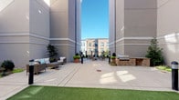 This amenity space combines sophisticated interior design with a functional outdoor courtyard, offering a well-rounded social environment. The outdoor area is framed by tall modern building facades finished in neutral gray tones, creating an enclosed yet open-air atmosphere. The spacious concrete patio is complemented by a patch of artificial turf, outdoor lounge furniture, potted greenery, and a brick-accented built-in gas grill station, making it well-suited for casual gatherings and outdoor entertaining.

Inside, the lounge and game room is thoughtfully designed with layered textures and refined finishes. Metallic and stone-effect wall treatments add visual depth throughout, while recessed lighting and stylish globe pendant fixtures set a warm, inviting tone. A full-size pool table anchors the game area, accompanied by a backgammon set and additional tabletop games on a central coffee table. A built-in wall unit houses a flat-screen television surrounded by organized shelving filled with books and decorative objects, illuminated by warm sconce lighting. Navy leather seating, green upholstered accent chairs, and bistro-style high-top tables complete the space. A wet bar with dark cabinetry and stainless steel appliances provides added convenience for entertaining. Glass-panel doors connect the interior directly to the outdoor courtyard.