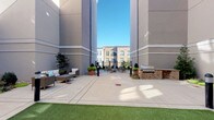 This amenity space combines sophisticated interior design with a functional outdoor courtyard, offering a well-rounded social environment. The outdoor area is framed by tall modern building facades finished in neutral gray tones, creating an enclosed yet open-air atmosphere. The spacious concrete patio is complemented by a patch of artificial turf, outdoor lounge furniture, potted greenery, and a brick-accented built-in gas grill station, making it well-suited for casual gatherings and outdoor entertaining.

Inside, the lounge and game room is thoughtfully designed with layered textures and refined finishes. Metallic and stone-effect wall treatments add visual depth throughout, while recessed lighting and stylish globe pendant fixtures set a warm, inviting tone. A full-size pool table anchors the game area, accompanied by a backgammon set and additional tabletop games on a central coffee table. A built-in wall unit houses a flat-screen television surrounded by organized shelving filled with books and decorative objects, illuminated by warm sconce lighting. Navy leather seating, green upholstered accent chairs, and bistro-style high-top tables complete the space. A wet bar with dark cabinetry and stainless steel appliances provides added convenience for entertaining. Glass-panel doors connect the interior directly to the outdoor courtyard.