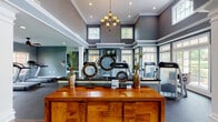 This upscale fitness center combines refined architectural design with a comprehensive suite of professional-grade exercise equipment. The main workout area commands attention with its dramatically high ceilings, crown molding, and white-trimmed clerestory windows that flood the space with natural light. Elegant chandelier lighting adds a distinctive, polished ambiance rarely found in fitness facilities, while decorative columns frame the open floor plan and contribute to the space's sophisticated character.

The main strength training area is outfitted with a full array of Cybex resistance machines positioned on durable rubber flooring, providing ample room to move between stations. Large multi-pane windows along the perimeter offer views of surrounding greenery, creating a bright and energizing environment. A stylish reception or transition area features a rich hardwood credenza with decorative accents, serving as a welcoming focal point.

A secondary cardio room, finished with warm hardwood flooring, houses Cybex ellipticals and upright bikes alongside a ceiling fan and an arched window overlooking the outdoors. An additional room with hardwood floors provides treadmills and open space suited for stretching or light activity. Throughout the facility, the cool gray and slate-blue color palette, recessed lighting, and thoughtful décor combine to create a motivating yet refined workout environment.