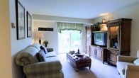 This single-floor residence offers a comfortable and elegantly appointed living environment with a timeless, traditional aesthetic. The living room serves as the heart of the space, showcasing plush, patterned upholstered seating arranged around a tufted leather ottoman that doubles as a central coffee table — a refined focal point for the room. A large, ornate wooden entertainment armoire houses a flat-screen television and provides generous storage, adding a rich, classic character to the space. Soft, neutral-toned carpeting extends throughout, contributing to a warm and inviting ambiance. A sliding glass door floods the room with natural light and offers direct access to an outdoor area, where greenery is visible beyond, creating a pleasant connection between interior and exterior. Decorative window treatments in a striped green fabric frame the door with a tailored elegance. Accent lighting from table lamps casts a soft, warm glow, complementing the natural light and enhancing the cozy atmosphere. The clean white walls and smooth ceilings provide a bright, airy backdrop that allows the furnishings and décor to take center stage throughout this thoughtfully designed single-level home.