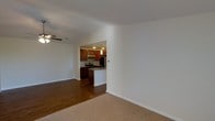 This single-floor residence offers a clean, open-concept layout that seamlessly connects the living area to the kitchen. The living space is finished with warm, neutral-toned carpet and crisp white walls, creating a bright and welcoming atmosphere. A ceiling fan adds both functional airflow and a subtle design element to the room.

The kitchen is accessible through an open arched passageway and features rich, dark wood cabinetry that provides ample storage. Stainless steel appliances — including a refrigerator, microwave, and range — lend a modern, polished look to the cooking space. A peninsula with a stone-style countertop offers additional prep space and casual seating potential. Pendant lighting above the peninsula adds a warm, focused glow that enhances the kitchen's inviting character.

The hardwood flooring in the kitchen contrasts nicely with the carpeted living area, helping to visually define the two spaces while maintaining a cohesive overall aesthetic. The neutral color palette throughout keeps the space feeling airy and versatile. Overall, the layout is practical and well-appointed, making this a comfortable and functional single-floor living space.