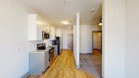 This single-floor residence offers a thoughtfully designed layout with contemporary finishes throughout. The galley-style kitchen is a standout feature, equipped with stainless steel appliances including a refrigerator, electric range, and over-the-range microwave. Gray lower cabinetry is paired with white upper cabinets, some open shelving, and a built-in wine rack, all complemented by granite countertops and a classic subway tile backsplash. Light wood-style flooring runs through the kitchen and main living areas, creating a warm, cohesive aesthetic. The living space benefits from natural light streaming through double French doors that open to an outdoor area, with a ceiling fan overhead adding to the comfort of the room. The bedroom features soft carpeting, a ceiling fan, and direct access to a spacious walk-in closet with built-in shelving. The full bathroom is cleanly finished with a granite-topped gray vanity, a large mirror, a combination tub and shower, and matching wood-style tile flooring. The overall design maintains a neutral, modern palette with gray accents and clean white walls, offering a bright and welcoming ambiance throughout.