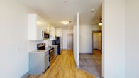 This single-floor residence offers a thoughtfully designed layout with contemporary finishes throughout. The galley-style kitchen is a standout feature, equipped with stainless steel appliances including a refrigerator, electric range, and over-the-range microwave. Gray lower cabinetry is paired with white upper cabinets, some open shelving, and a built-in wine rack, all complemented by granite countertops and a classic subway tile backsplash. Light wood-style flooring runs through the kitchen and main living areas, creating a warm, cohesive aesthetic. The living space benefits from natural light streaming through double French doors that open to an outdoor area, with a ceiling fan overhead adding to the comfort of the room. The bedroom features soft carpeting, a ceiling fan, and direct access to a spacious walk-in closet with built-in shelving. The full bathroom is cleanly finished with a granite-topped gray vanity, a large mirror, a combination tub and shower, and matching wood-style tile flooring. The overall design maintains a neutral, modern palette with gray accents and clean white walls, offering a bright and welcoming ambiance throughout.