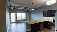 This single-floor space showcases a thoughtfully designed open-concept layout that seamlessly blends the kitchen and living areas. The kitchen is a standout feature, centered around a large granite-topped island with an undermount sink, elegant pendant lighting with industrial-style fixtures, and dark espresso cabinetry paired with stainless steel appliances. Exposed ductwork running along the ceiling adds a distinctive industrial character to the space, complementing the modern aesthetic. Rich hardwood flooring flows throughout, grounding the cool blue-grey walls and creating a warm contrast against the darker cabinetry. The living area benefits from generous natural light streaming through large windows, creating a bright and welcoming atmosphere. A full-light interior door provides access to additional areas of the unit. The open floor plan promotes a sense of spaciousness and ease of movement between living and cooking areas, making it ideal for both everyday living and entertaining. The combination of industrial accents, quality finishes, and natural light gives this space a contemporary yet inviting character.