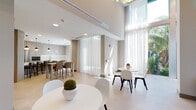 Located at 750 NW 43rd Ave in Miami, FL, this contemporary amenity space offers a sophisticated and open environment designed for comfort and social interaction. The single-floor layout is distinguished by impressively soaring double-height ceilings that lend an airy, expansive feel throughout.

Floor-to-ceiling sheer curtains line the perimeter, filtering abundant natural light while framing lush tropical greenery visible just beyond the glass — a subtle nod to the South Florida surroundings. The light-toned large-format tile flooring runs seamlessly across the space, unifying each area.

Multiple distinct seating zones are thoughtfully arranged to accommodate a variety of uses. Casual lounge areas feature plush leather sofas and accent chairs positioned around a built-in wall unit with lit display shelving, decorative art objects, and a central media screen. Dining and bar-height tables with upholstered seating provide flexible spaces for both individual use and group gatherings.

The fully equipped kitchen area includes stainless steel appliances, ample cabinetry in a warm wood-tone finish, a kitchen island with bar seating, and pendant lighting overhead. Colorful artwork accents the walls, adding vibrancy to the otherwise neutral, modern palette. A sputnik-style chandelier serves as a striking focal point, completing the refined yet approachable aesthetic of this well-appointed communal space.