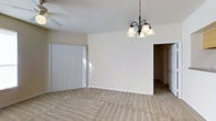 This single-floor residential space is designed with simplicity and functionality in mind, offering a comfortable and versatile layout. The interior is finished in soft, neutral tones throughout, creating a bright and welcoming ambiance that complements a variety of décor styles.

The main living and dining area is notably open, featuring plush wall-to-wall carpeting in a warm beige hue that adds texture and comfort underfoot. An elegant multi-bulb chandelier with frosted glass shades serves as a focal point overhead, providing warm ambient lighting to the dining or gathering space. A ceiling fan is also present, adding practical air circulation to the room.

Natural light enters through a large sliding door or window fitted with vertical blinds, allowing for adjustable brightness and privacy. The transition from carpet to hard surface flooring marks the passage into an adjoining hallway or room, suggesting a thoughtfully segmented floor plan.

The walls are painted in a clean, neutral off-white, contributing to the overall sense of openness and light. White baseboards frame the room neatly, adding a crisp finishing detail. The space flows naturally between areas, making it well-suited for everyday living. Overall, this home offers a clean, move-in-ready environment with classic residential finishes.