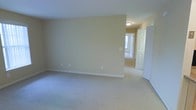 This single-floor space presents a straightforward, functional layout with a calm, neutral aesthetic throughout. The main room is generously sized, featuring wall-to-wall carpeting in a light gray tone that adds warmth underfoot while complementing the soft beige walls. The room is open and unfurnished, allowing the full sense of its spaciousness to be appreciated. White baseboards provide a clean contrast against the walls, and standard electrical outlets are positioned at convenient intervals. A flush-mount ceiling light fixture provides overhead illumination. An open doorway leads to an adjacent room, also carpeted, which appears to receive natural light from a window, suggesting a bright and airy feel in that area. A white paneled door is visible nearby, likely leading to a closet or additional room, offering practical storage or access options. The overall palette — warm beige walls, light carpet, and white trim — creates a cohesive, neutral environment that is versatile and easy to personalize. The space presents a clean, move-in-ready condition with a simple, uncluttered character suited to a variety of uses.