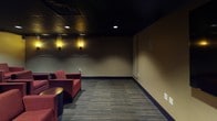 This single-floor screening and lounge space is thoughtfully designed to create an intimate, theater-like atmosphere. The room is anchored by a large wall-mounted flat-screen television positioned prominently on one wall, flanked by in-wall speakers and acoustic panels that enhance audio performance throughout the space.

The dark, tiled drop ceiling is outfitted with recessed downlights that cast a focused, subdued glow across the room, contributing to a cinematic ambiance. Warm amber wall sconces on the rear wall add layered lighting, softening the overall mood while maintaining visibility.

Seating consists of multiple plush red upholstered armchairs arranged in groupings throughout the room, offering flexible configurations for both small and larger gatherings. Neutral-toned sofa seating is also present, providing additional comfort. Small side tables are interspersed among the chairs for convenience.

The flooring features multi-toned striped carpeting that runs throughout the space, adding texture underfoot while absorbing sound. Warm tan walls, dark blue baseboards, and framed artwork on the walls complete the polished, lounge-style aesthetic. A single door with a glass panel provides access, with an illuminated exit sign overhead reinforcing the venue-style character of the space.