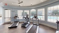 This fitness center is thoughtfully designed to accommodate a wide range of workout needs within a single, open-plan floor. The space is divided into distinct functional zones, including a dedicated Training Zone stocked with TRX suspension systems, resistance bands, medicine balls, and stability equipment. A comprehensive free weights area features a full dumbbell rack alongside multiple strength machines, while a cable crossover station anchors the central floor space.

A standout architectural feature is the curved cardio alcove, characterized by a rounded wall of large arched windows that flood the area with natural light and offer outdoor views of what appears to be a pool. This section houses treadmills, ellipticals, and stair climbers from brands including True Fitness. Ceiling fans are installed throughout, complementing recessed lighting to maintain airflow and comfort.

The interior design reflects a refined aesthetic, with pale blue walls, white crown molding, coffered ceilings, and decorative lattice-framed mirrors that visually expand the space. Dark hardwood-style flooring runs throughout, and glass doors provide direct outdoor access, adding to the sense of openness and connectivity to the surrounding amenities.
