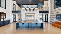 Located at 1000 North 44th Street in Phoenix, Arizona, Hangar 44 is a thoughtfully designed, single-floor community clubhouse that draws inspiration from aviation heritage. The space makes a striking first impression with its grand entrance lobby, featuring soaring double-height ceilings, floor-to-ceiling glass windows, and a warm wood-accented entry door that floods the interior with natural light and outdoor views of the surrounding Phoenix skyline.

At the heart of the space is a central work and gathering area anchored by a large dark stone communal table with modern blue bar seating, accented by an eye-catching cluster of oversized pendant lights overhead. The prominent Hangar 44 branded wall feature adds a distinctive industrial character throughout.

The resident lounge area is expansive and light-filled, with dramatic curtain-wall glazing opening to outdoor terrace views. Sleek leather and metallic lounge chairs, plush sofas, a fireplace, and built-in shelving curated with aviation-themed décor create a comfortable yet sophisticated atmosphere.

A fully equipped bar and dining area features green tile backsplash, open bottle shelving, and bar seating, complemented by a resident kitchen with ample counter space and appliances. Polished concrete flooring, exposed structural trusses, and bold dark steel accents tie the entire space together with an industrial-modern aesthetic.