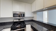 This single-floor space offers a thoughtfully designed kitchen that serves as a functional and visually appealing centerpiece. The kitchen is outfitted with stainless steel appliances, including an over-the-range microwave and a gas range, both contributing a sleek, modern aesthetic. Dark granite countertops with subtle flecks of light provide a striking contrast against the crisp, white shaker-style cabinetry that lines the walls from counter to ceiling, maximizing storage space throughout. The cabinetry features a clean, classic design with recessed panel doors, lending the kitchen a timeless and polished look. The L-shaped countertop layout makes efficient use of the available space, offering ample room for food preparation. The light, neutral color palette of the white cabinets and walls creates a bright and welcoming atmosphere. Overall, the kitchen is designed with both practicality and style in mind, combining durable materials with a refined aesthetic that would appeal to a wide range of occupants. The single-floor layout of the space ensures convenient and accessible living throughout.