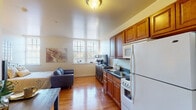 Located at 1833 N Third St in Harrisburg, Pennsylvania, this single-floor unit offers a well-organized, open-concept layout that efficiently combines living, sleeping, and cooking spaces. Warm hardwood flooring runs throughout the main areas, lending a cohesive and inviting feel to the entire space.

The kitchen is equipped with rich, warm-toned wood cabinetry — seen in varying finishes across the unit — and includes a full suite of white appliances: a refrigerator, electric range, and over-the-range microwave. A double sink with dark countertops completes the functional workspace, which flows naturally into the living and dining areas.

The open living area accommodates both a seating arrangement and a dining setup, with natural light provided by multiple windows featuring classic gridded panes. The bedroom area is generously sized, framed by two large windows that fill the room with abundant natural light and offer views of the surrounding neighborhood.

The bathroom is cleanly finished with tile flooring, a pedestal sink, toilet, and a walk-in shower enclosed by a curtain. Neutral wall tones and framed botanical artwork contribute to a calm, understated ambiance throughout the space.