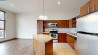 This thoughtfully designed single-floor unit offers a compact yet functional layout that maximizes every square foot of its living space. The open-concept kitchen and dining area serves as the heart of the unit, showcasing rich warm-toned wood cabinetry that extends floor to ceiling, a classic white subway tile backsplash, and a full suite of stainless steel appliances including a refrigerator, electric range, over-the-range microwave, and dishwasher. A curved granite-topped kitchen island provides additional prep space and a natural divider between the kitchen and dining area. Light tile flooring in the kitchen transitions seamlessly into durable wood-look flooring throughout the rest of the unit. Soaring ceilings with an exposed ductwork detail lend an industrial-modern character to the space, while a stylish pendant light anchors the kitchen area. Large floor-to-ceiling windows flood the living area with natural light and frame expansive urban views of the surrounding cityscape. The bathroom is clean and well-appointed, featuring a wood-toned vanity with a stone countertop, an integrated sink, and a full tub-shower combination with accessibility grab bars. The unit is completed by an in-unit laundry area, adding everyday convenience to this modern urban residence.