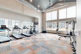 Located at 16631 E Colfax Ave in Aurora, CO, this fitness center is a thoughtfully designed exercise space that offers a comprehensive workout environment. The room is defined by its dramatic vaulted ceiling with a dark painted upper section, exposed ductwork, and ceiling fans that lend an industrial-modern aesthetic. Large windows along multiple walls flood the interior with natural light and provide open views of the surrounding neighborhood.

The exercise room is outfitted with a full lineup of cardio equipment, including multiple treadmills, an elliptical trainer, and a stationary bike. A cable resistance machine and a dedicated dumbbell rack with a wide range of free weights support strength training, while a wall-mounted mirror expands the sense of space and allows for form-checking during workouts. A well-stocked functional training station features kettlebells, medicine balls, stability balls, and exercise mats for dynamic training routines.

The floor is covered in a distinctive multi-toned carpet tile in shades of grey and orange that adds color and comfort underfoot. A flat-screen TV is mounted on the wall for entertainment during workouts. A glass-paneled door leads to an adjacent Kids Room, making the facility family-friendly. Storage closets and a security camera round out the practical amenities of this well-appointed fitness center.