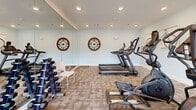 Located at 216–218 Campus Dr in Central, South Carolina, this fitness center is a thoughtfully designed exercise space that delivers a full workout experience in a compact, single-floor layout. The room features light gray walls, recessed ceiling lighting, and durable patterned carpet tiles that lend a clean, modern aesthetic throughout.

The space is outfitted with a comprehensive selection of cardio and strength equipment, including treadmills, elliptical trainers, a rowing machine, and a dumbbell rack with free weights. A wall-mounted flat-screen television adds entertainment for users during workouts. Decorative wall clocks provide both functional and visual interest along one wall.

A full-length mirror spans a significant portion of one wall, effectively amplifying the sense of space and allowing users to monitor their form. Large windows along the exterior wall flood the room with natural light and offer views of the surrounding grounds, while a glass-paneled entry door further enhances the bright, open feel. The combination of ample equipment, thoughtful décor, and abundant natural light makes this fitness center a practical and inviting amenity for residents of the Campus Drive community.