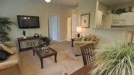 This single-floor residence centers around a living room finished in warm, neutral tones that create a calm and inviting atmosphere. The space is carpeted throughout, adding a soft, comfortable feel underfoot. The living area is furnished with a plush sofa accented by decorative throw pillows, a classic wooden coffee table with a glass top, and a matching accent chair, all arranged to encourage conversation and relaxation. A wall-mounted flat-screen television is positioned opposite the seating area, complemented by a console table below displaying decorative accents, including a traditional mantel clock. A framed art piece adorns one wall, while a table lamp provides warm ambient lighting. The open layout allows clear sightlines to interior doorways leading to additional rooms, suggesting a practical and connected floor plan. A pass-through or shelf detail near one wall hints at a nearby kitchen or dining area. The overall design aesthetic leans traditional, with dark wood furniture contrasting against the light-colored walls and carpet, creating a cohesive and well-appointed interior that balances functionality with classic style.