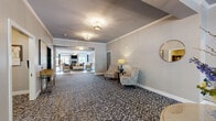 Located at 32 Aviation Rd in Albany, NY, this single-floor community space offers a generous and thoughtfully designed layout that balances comfort with functionality. The expansive living room serves as the heart of the space, anchored by a striking stone fireplace with a mounted television, surrounded by plush blue seating arranged on a patterned area rug. Large glass doors flood the room with natural light and provide access to an outdoor patio, enhancing the open, airy atmosphere. Tray ceilings with crown molding add an architectural elegance throughout. An intimate lounge area off the hallway features warm sconce lighting, tufted seating, and refined décor, creating a welcoming retreat. The combined dining room presents a bright, naturally lit setting with large windows, wood-look flooring, and a round dining table with upholstered chairs. The kitchen is outfitted with rich dark cabinetry, stainless steel appliances including a range, microwave, and refrigerator, complemented by white tile backsplash and light countertops. Consistent wood-look flooring flows throughout the entire space, unifying each area with a warm, cohesive aesthetic. The overall design reflects a polished, residential character well-suited for communal gatherings.