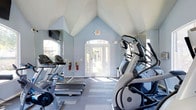 This single-story fitness center offers a bright and welcoming workout environment designed to support an active lifestyle. The space is defined by its striking vaulted ceiling with angular, geometric details finished in white, which draws the eye upward and creates a sense of openness. Soft blue walls contribute to a calm, energizing atmosphere throughout the room.

Natural light flows generously into the space through arched windows and a set of glass-paneled double doors at the entrance, offering views of the outdoors and creating an airy feel. The flooring features a striped, multi-toned pattern that adds visual interest while providing a practical surface for exercise.

The fitness center is equipped with a row of Matrix-brand treadmills and at least one Matrix elliptical machine, catering to a range of cardio workouts. Wall-mounted mirrors on multiple sides enhance the sense of space and allow users to monitor their form during exercise. A mounted television is also present, adding an entertainment option for longer workout sessions. A fire extinguisher and clearly marked exit signage reflect attention to safety throughout the facility.